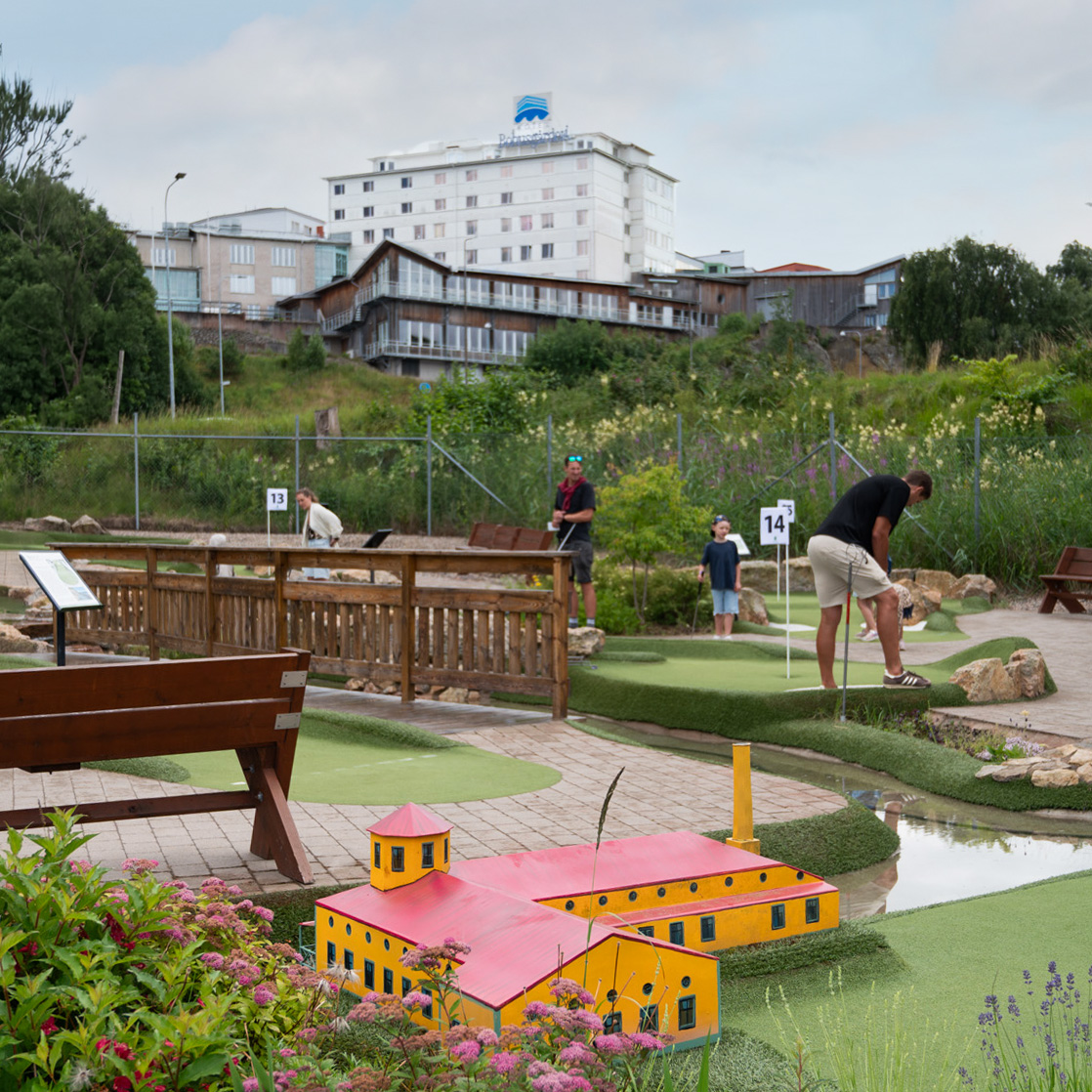 Äventyrsgolfbanan med en gul byggnad i miniatyr, spelande besökare och Bohusgården på höjden i bakgrunden.